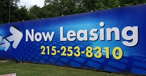 A 13oz oversized vinyl banner on display outdoors reading "Now Leasing" with a phone number printed below.