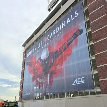 Oversized 9 oz heavy duty mesh banner on the side of a large building, printed with a promotion for the Louisville Cardinals.